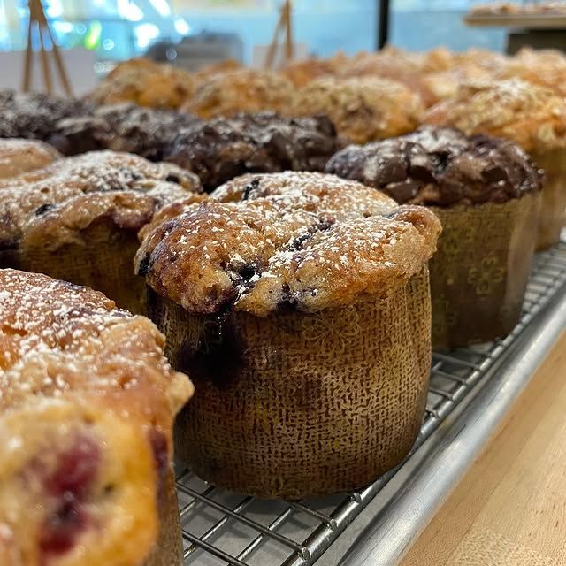 A close-up of various baked muffins dusted with powdered sugar, lined up on a metal cooling rack in a bakery.