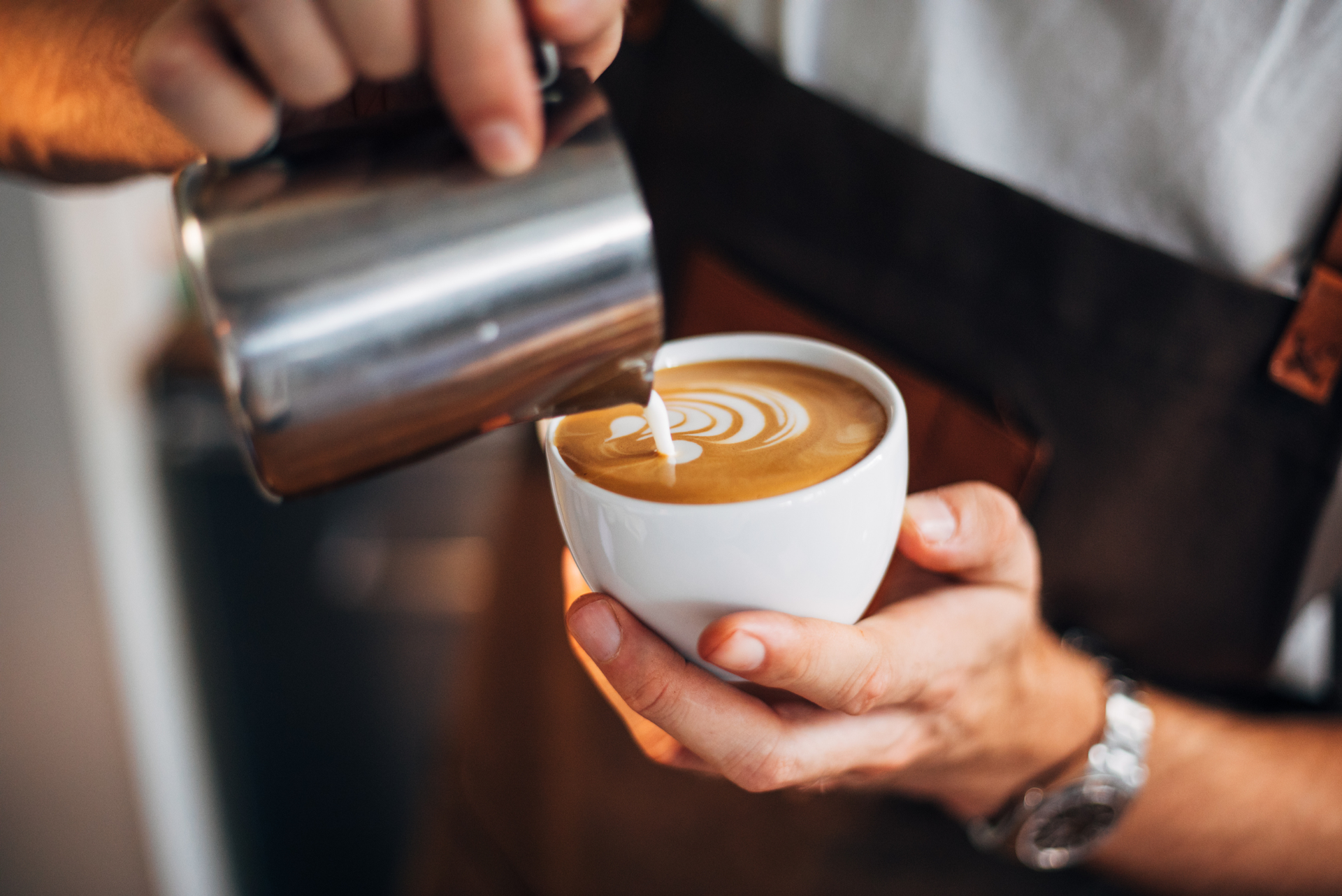 A barista pours steamed milk from a metal pitcher into a white cup to create latte art.