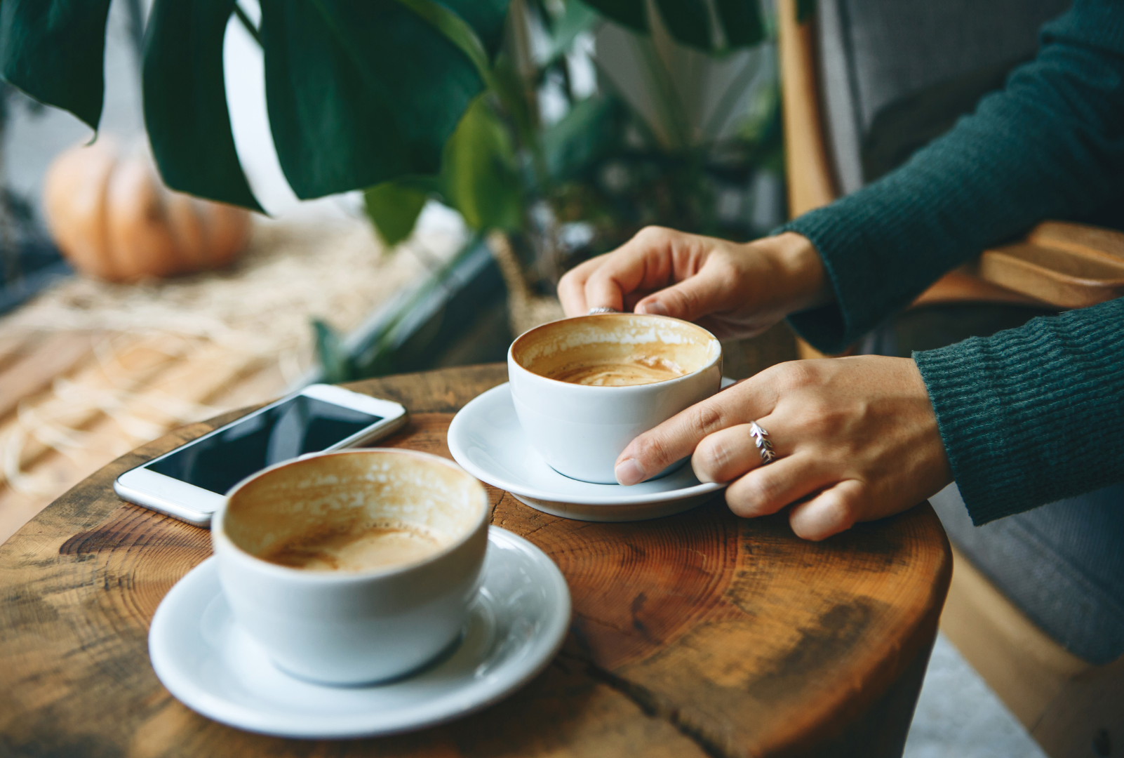 A person’s hands rest on a wooden table near two coffee cups and a smartphone, with a green plant in the background.