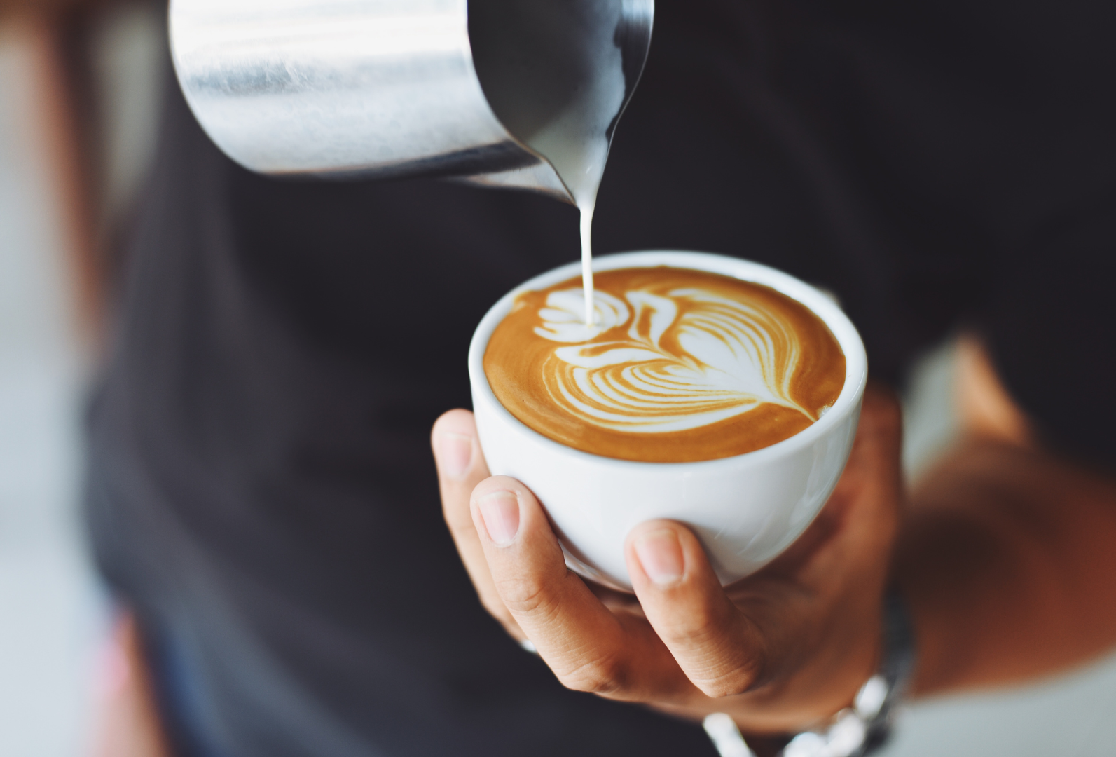 A person pouring steamed milk from a metal pitcher into a cup of coffee, creating a delicate rosetta latte art design.