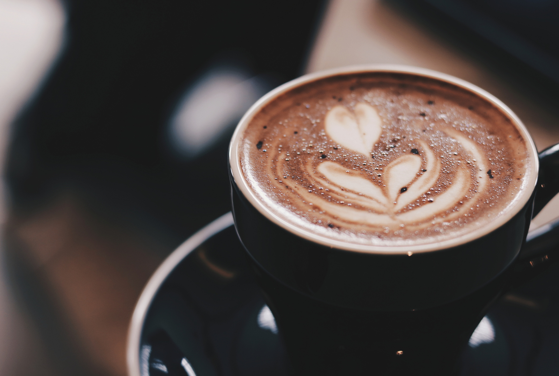 A close-up, high-angle shot of a dark ceramic cup filled with latte art foam, resting on a matching saucer.