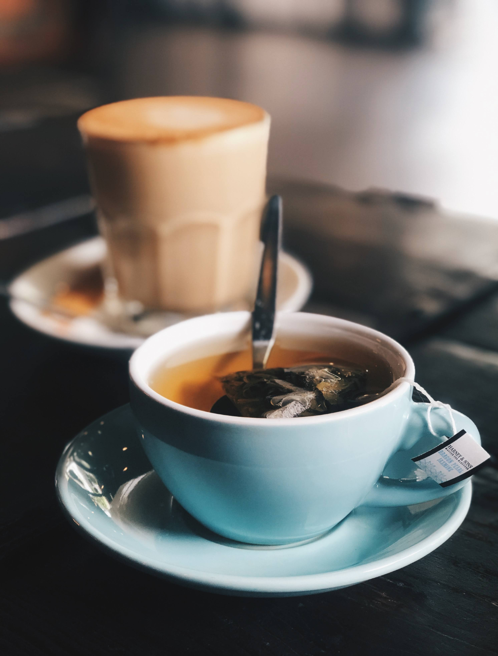 A light blue cup of tea with a tea bag inside sits on a saucer next to a blurred glass of latte on a dark wooden table.