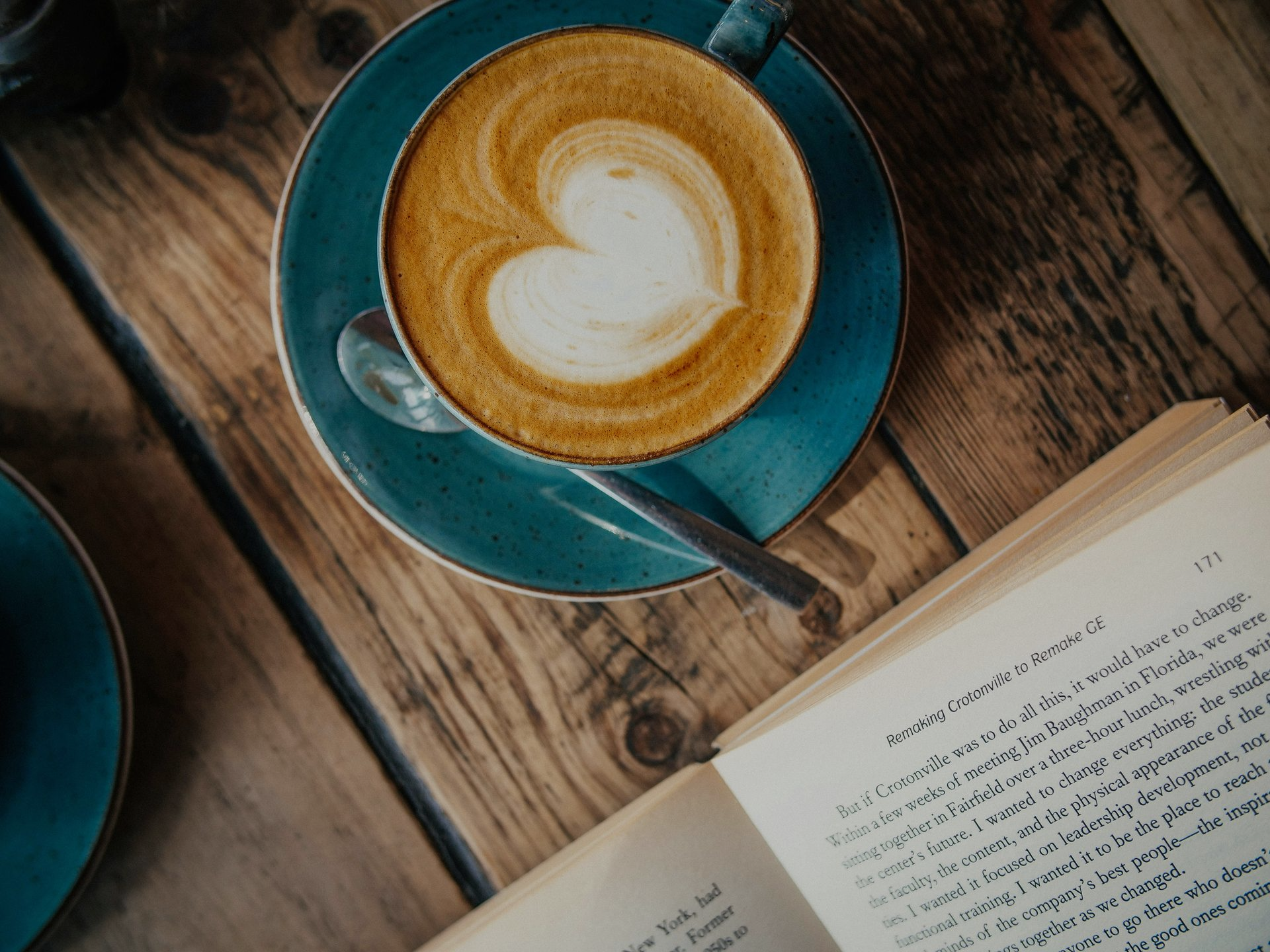 A latte with a heart-shaped foam design sits on a blue saucer next to an open book on a rustic wooden table.