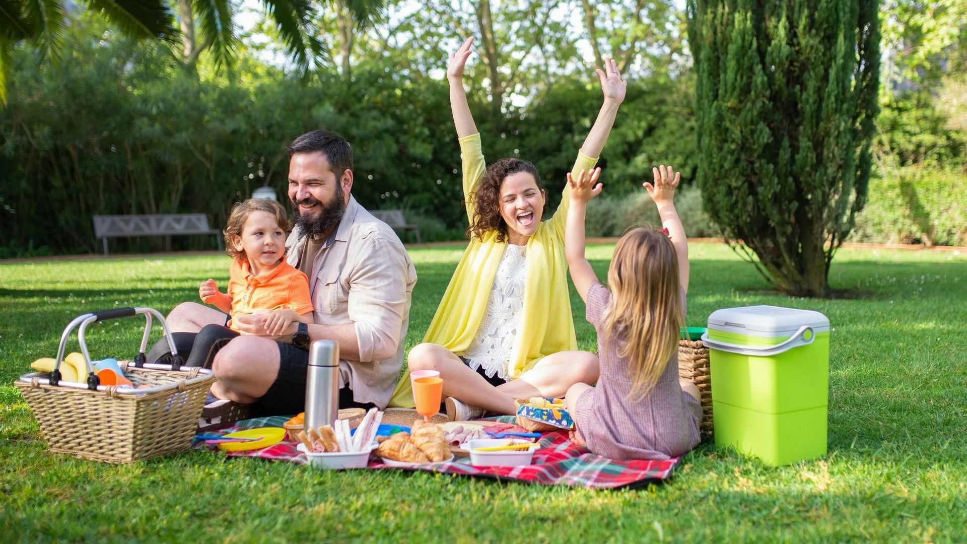 A family is sitting on a blanket in the park. They are having a picnic, and are smiling.