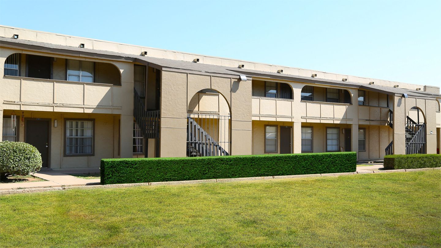 Two-story apartment building with beige facade, arched accents, and green hedges.