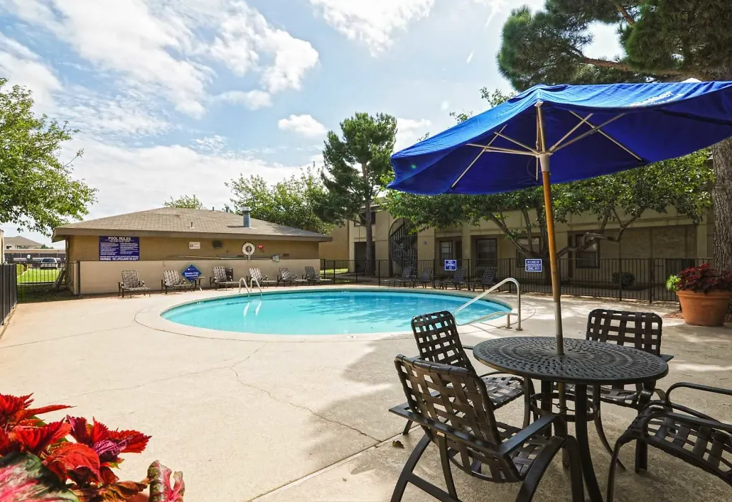 Pool area with blue umbrella, table and chairs, and building in the background. Sunny day.