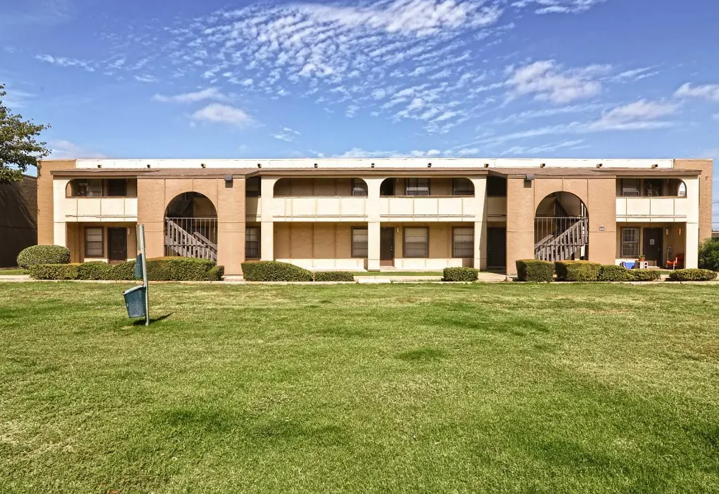 Two-story brick apartment building with arched entryways, green lawn, and blue sky.