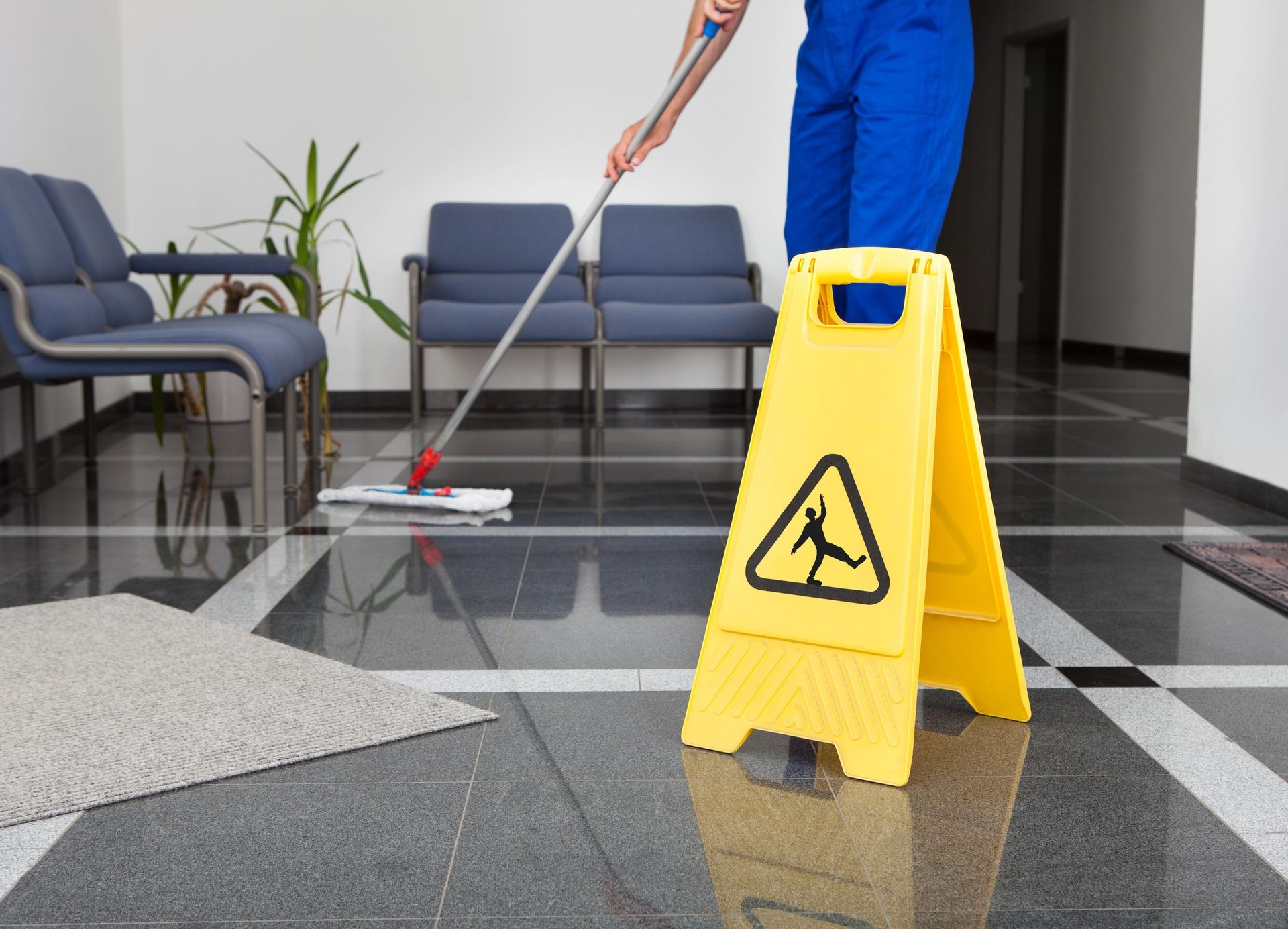 Una persona sta lavando un pavimento bagnato accanto a un cartello giallo di avvertimento nella hall.