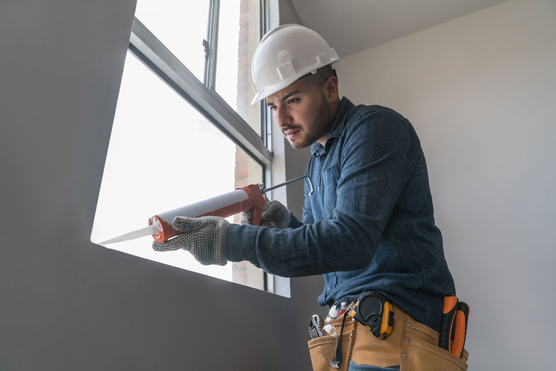 A man in a hard hat is applying sealant to a window.