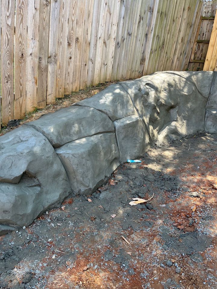 Rock retaining wall along a weathered wooden fence in a gravel yard, with a small toy near the base.