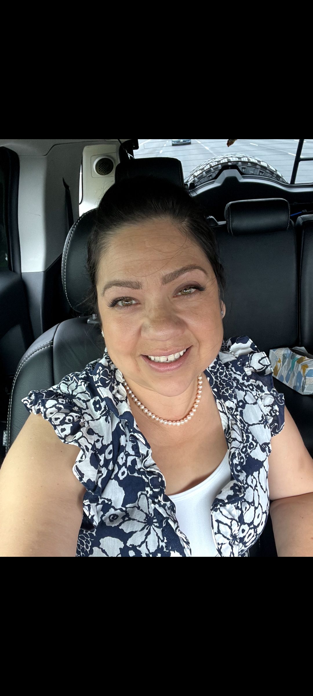 Woman smiling in a car. She has dark hair, and is wearing a patterned top and necklace.