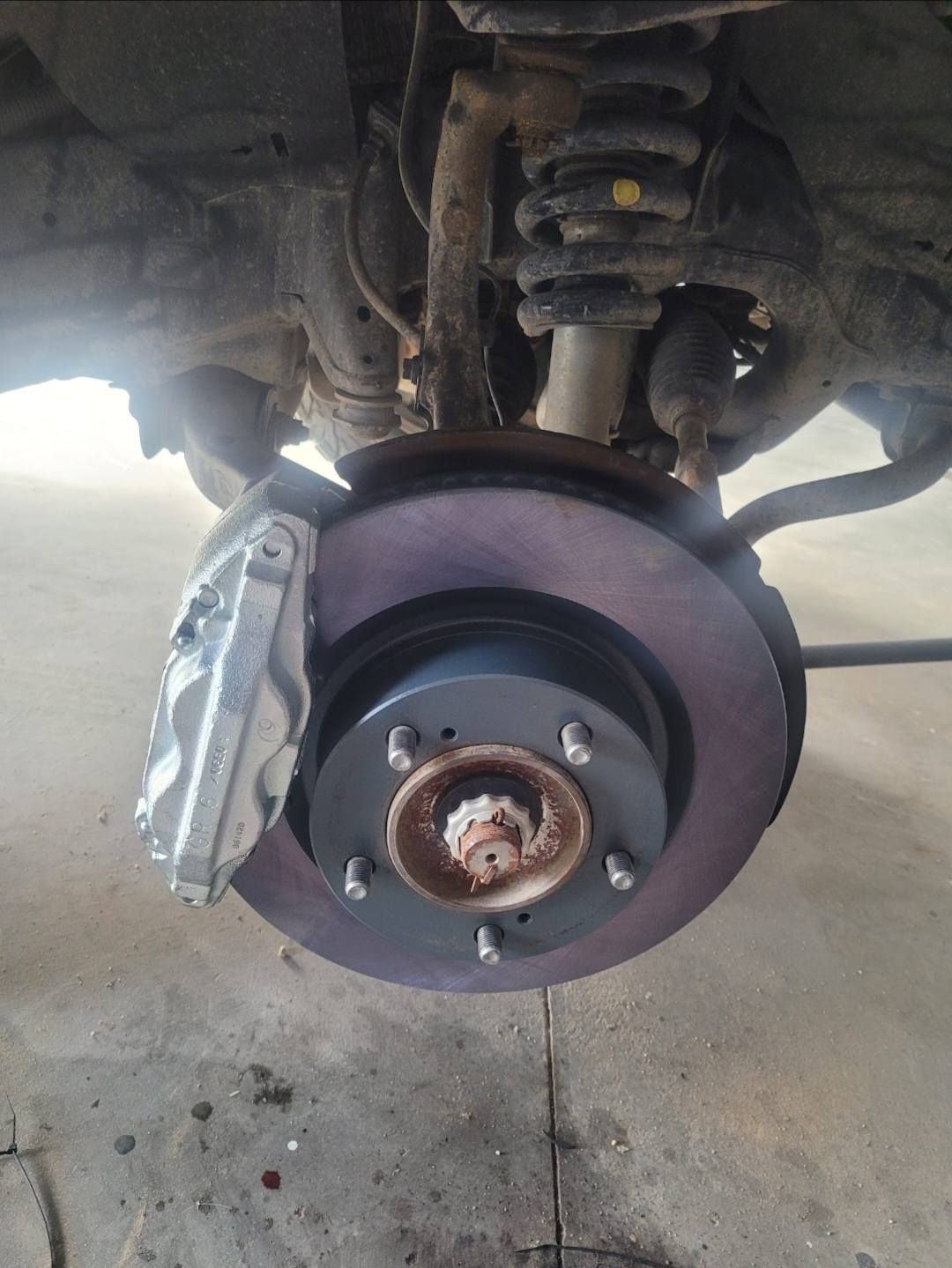 Car brake assembly, showing a rotor, caliper, and suspension components, with a rusty hub and visible brake dust.