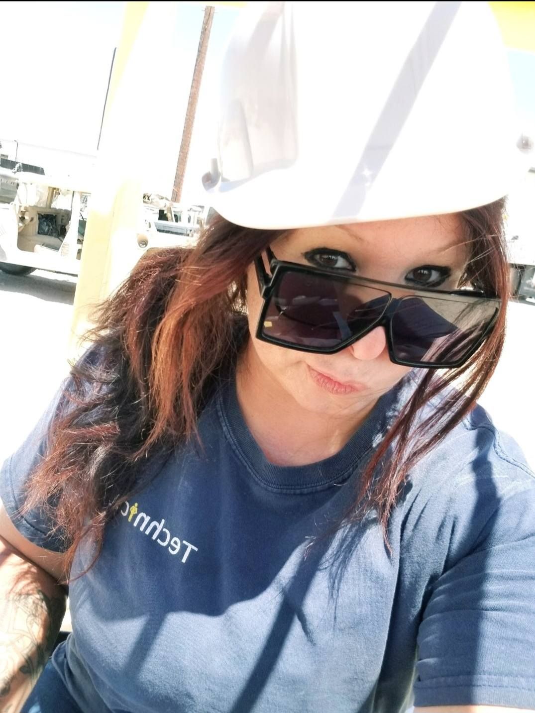 Woman in a white hard hat and sunglasses, wearing a blue t-shirt with text, outdoors in a sunny environment.