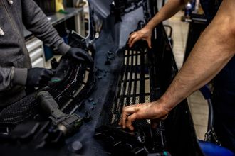 Two people working on a car bumper, one using a drill. Hands are visible, in a workshop setting.