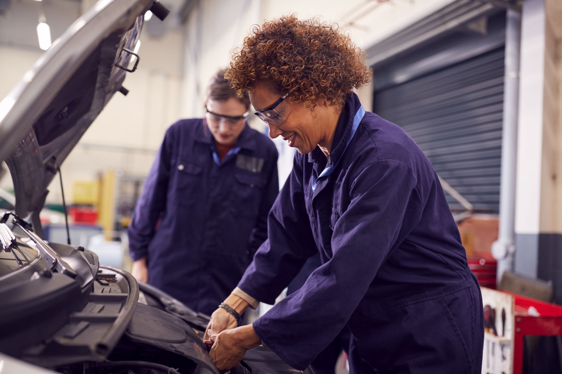 Two female mechanics working on a car engine in a garage, wearing safety glasses and blue coveralls.