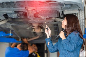 A mechanic inspects the underside of a car on a lift, using a tool to examine the vehicle. Other mechanics work in the background.