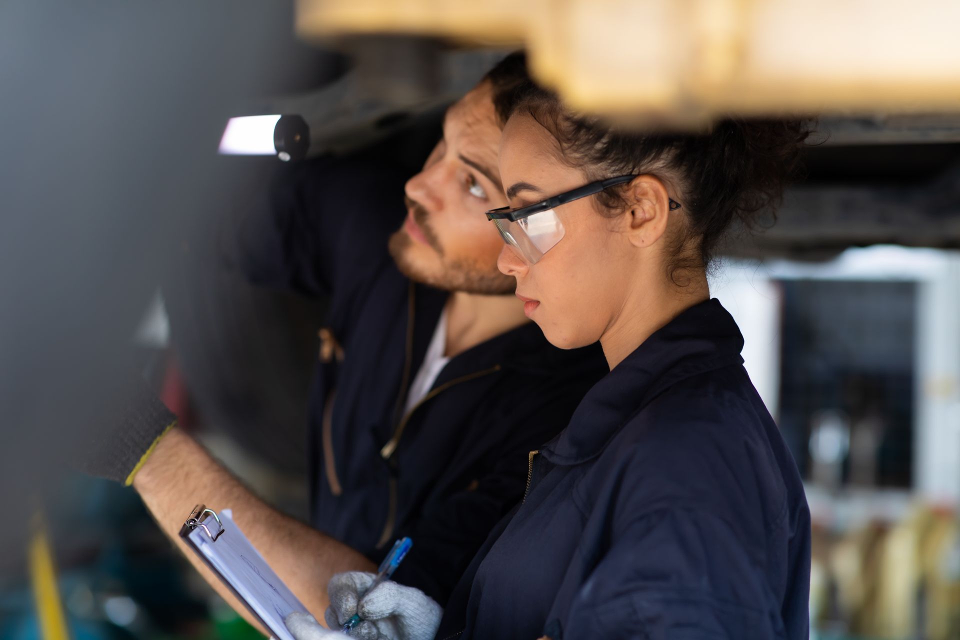 Two auto mechanics, a man and a woman, working under a car in a garage. The woman is wearing safety glasses and writing on a clipboard.