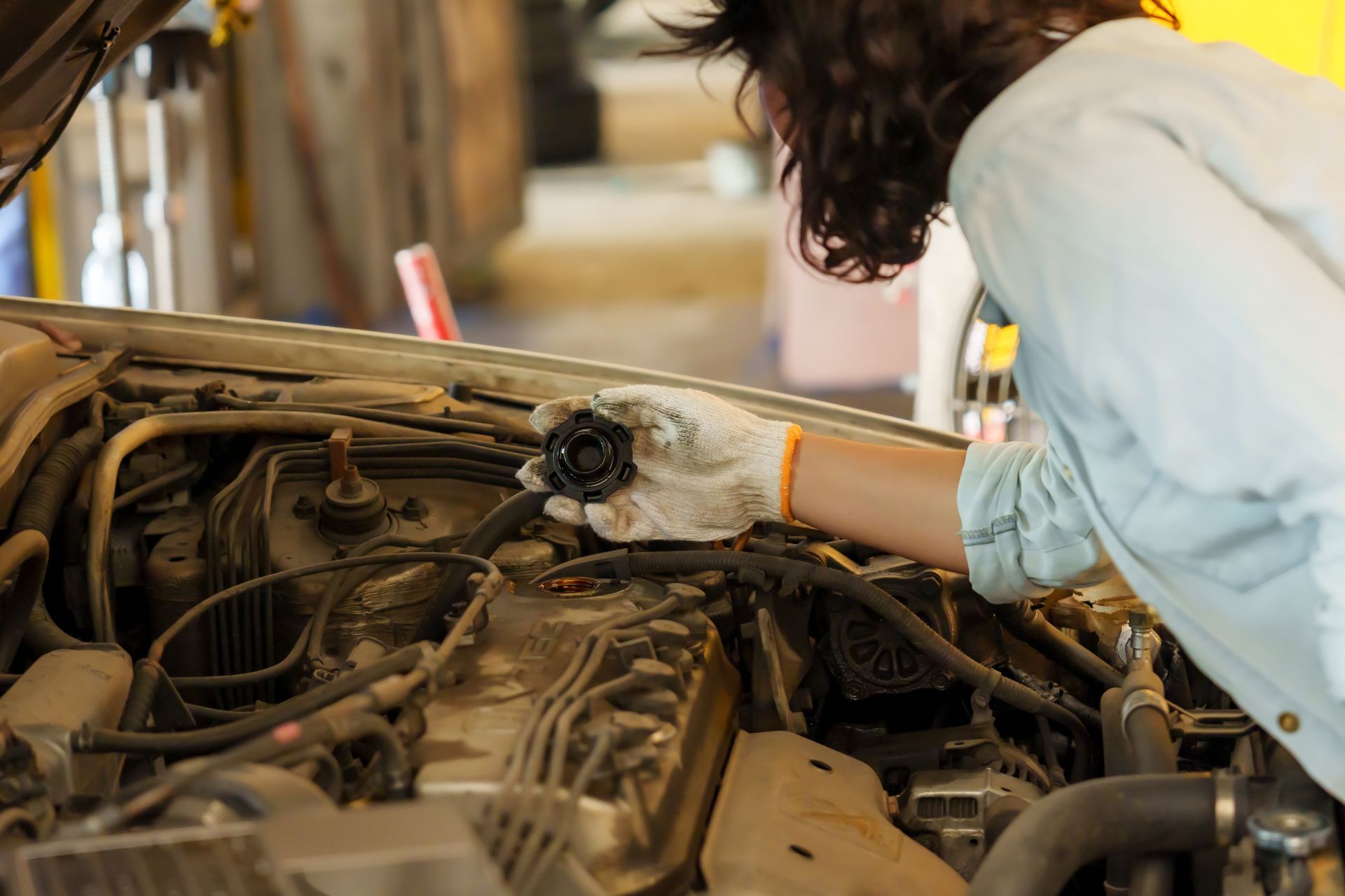 Woman in a garage inspecting a car engine, wearing gloves.