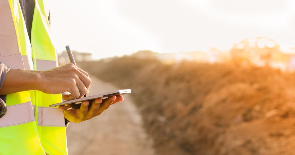 A Man In A Yellow Vest Is Writing On A Clipboard While Holding A Cell Phone — JTM Projects Australia Pty Ltd in Emerald, QLD