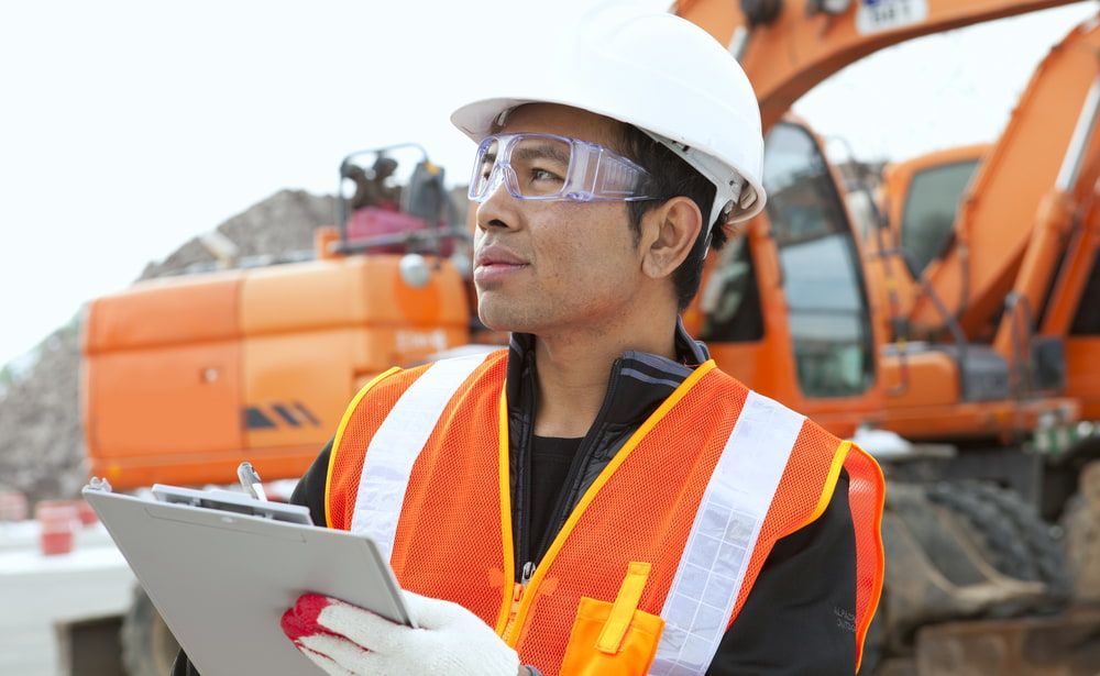 A Construction Worker Is Holding A Clipboard In Front Of An Excavator — JTM Projects Australia Pty Ltd in Mackay, QLD