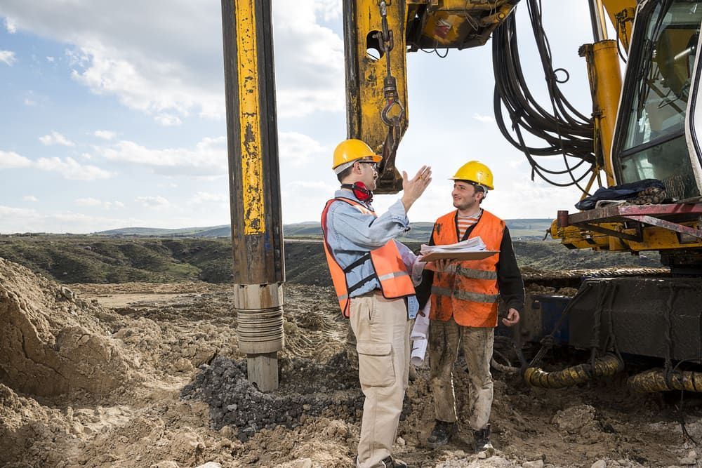 Two Construction Workers Are Standing In Front Of A Machine On A Construction Site — JTM Projects Australia Pty Ltd in Port Hedland, WA
