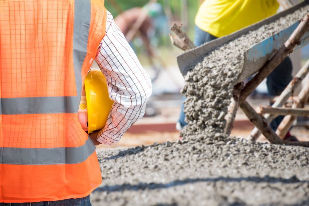 A Construction Worker Wearing A Hard Hat Is Standing In Front Of A Wheelbarrow Filled With Concrete — JTM Projects Australia Pty Ltd in Hollywell, QLD