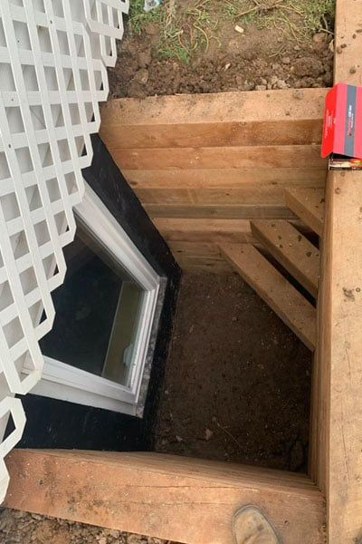 A window well constructed with wood and dirt. A white window is visible. A white lattice structure sits to the left.