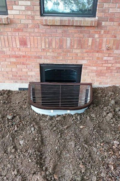 A brown window well cover sits in front of a basement window on a brick building. The well is surrounded by freshly turned soil.