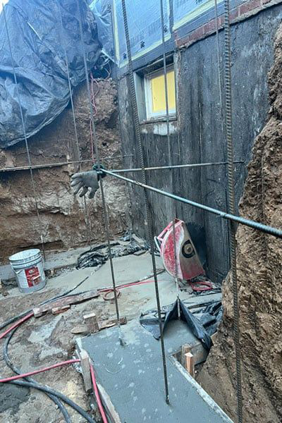 Construction site with a concrete foundation, rebar, and a saw. The wall behind it is dark, and there is a window with a yellow glow.