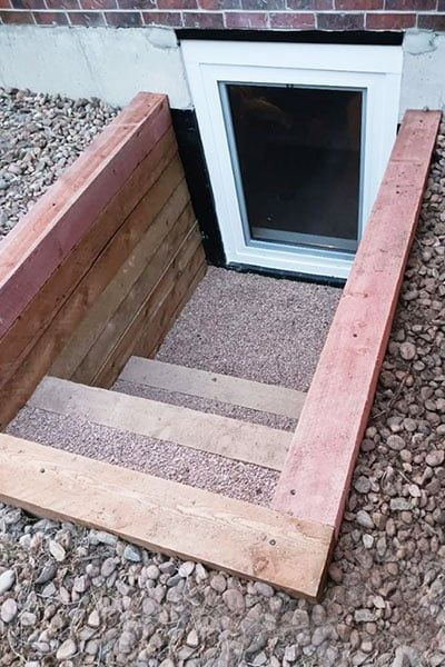 Window well with wooden steps leading to a basement window. Pinkish-brown wood frames the well, filled with gravel.