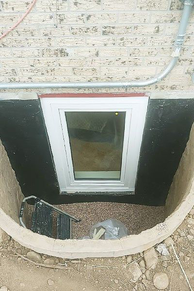Window well with a rectangular window. The well is lined with black waterproofing material and filled with gravel.