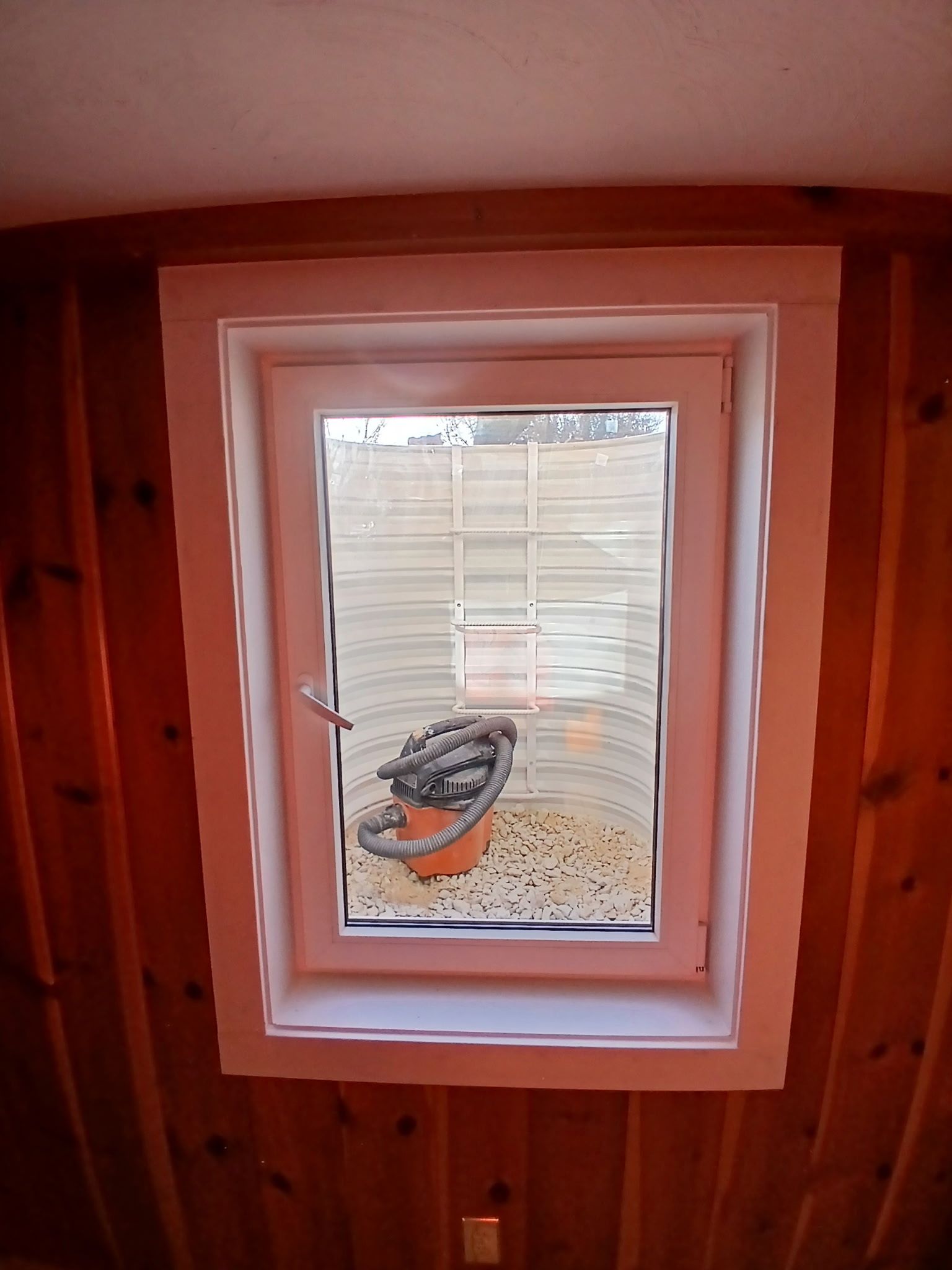 Living room with stone fireplace, large windows overlooking trees, and gray sofa. Wooden beams and neutral tones create a bright, natural atmosphere.