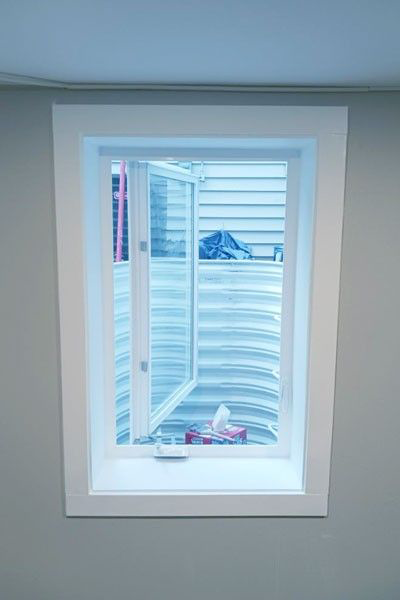 Open, white-framed window in a gray wall, revealing a basement window well. The window is partially open.