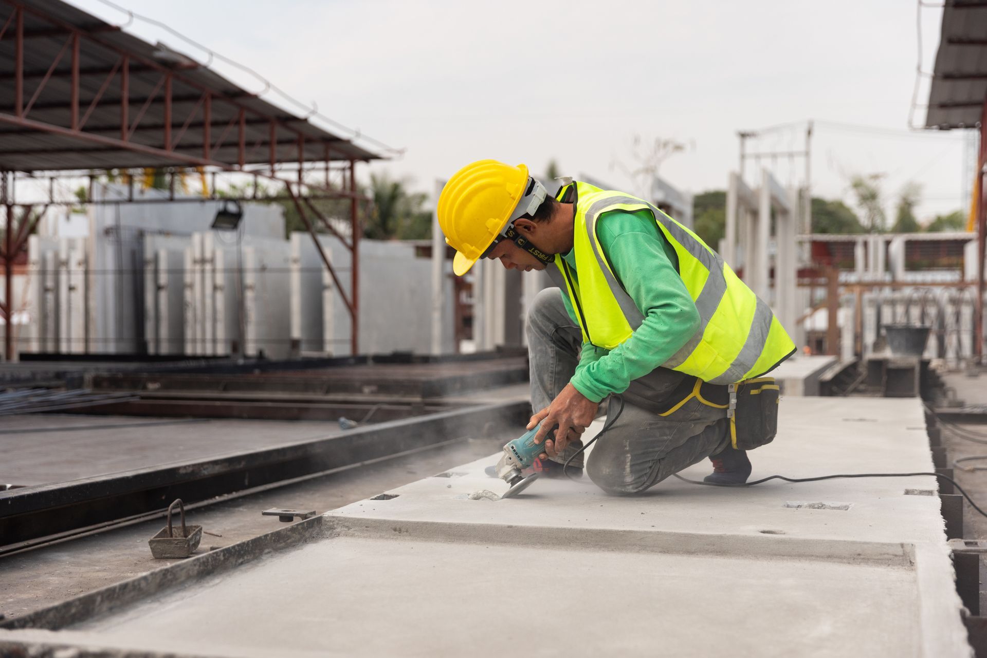 Construction worker wearing a yellow helmet and safety vest kneels, using a power saw to cut a concrete slab outdoors.