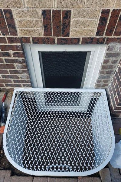 White security screen in front of a basement window with red and tan brick exterior.