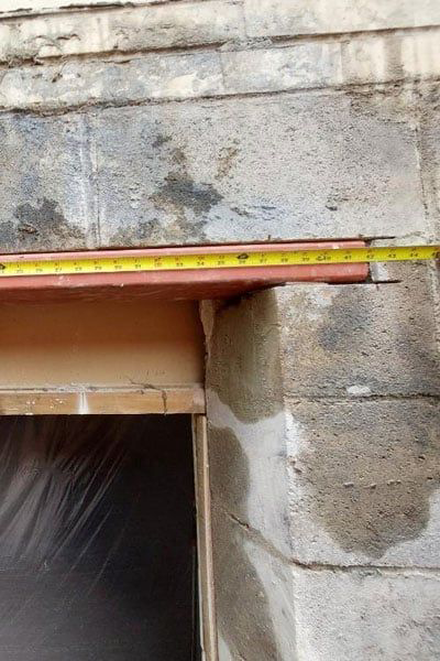 A measuring tape extends over a door frame. The frame is surrounded by aged concrete blocks. The opening is covered with plastic.