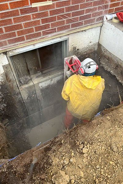 Person in yellow protective gear cutting concrete around a basement window. The setting is outdoors, beside a brick building.