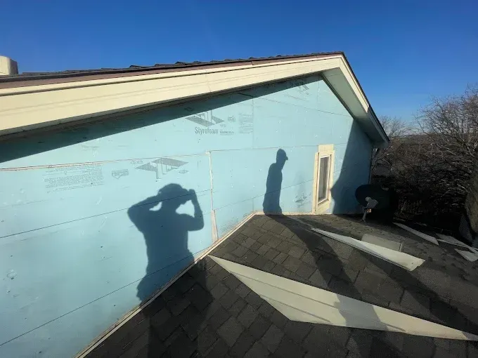 A person's shadow on blue house wrap; dark roof, light blue sky.