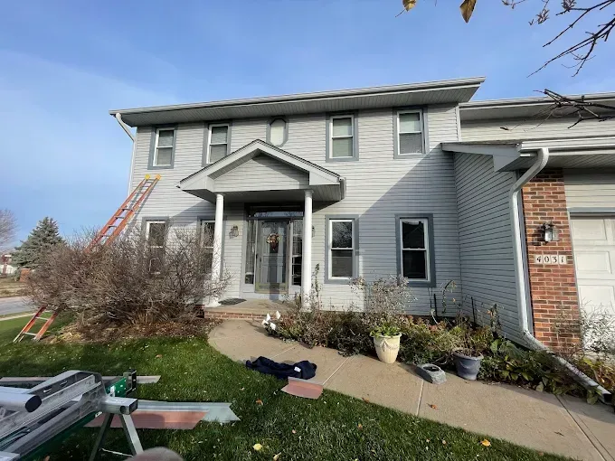 Two-story gray house with a brick garage. A ladder leans against the house; a pathway leads to the front door.