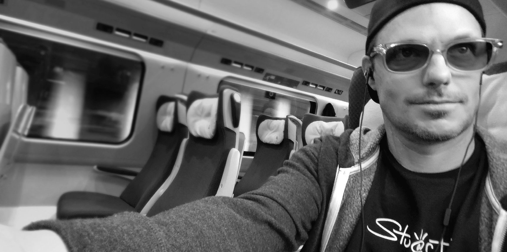 A person wearing sunglasses and a cap sits on a train, taking a selfie with empty seats visible in the background.