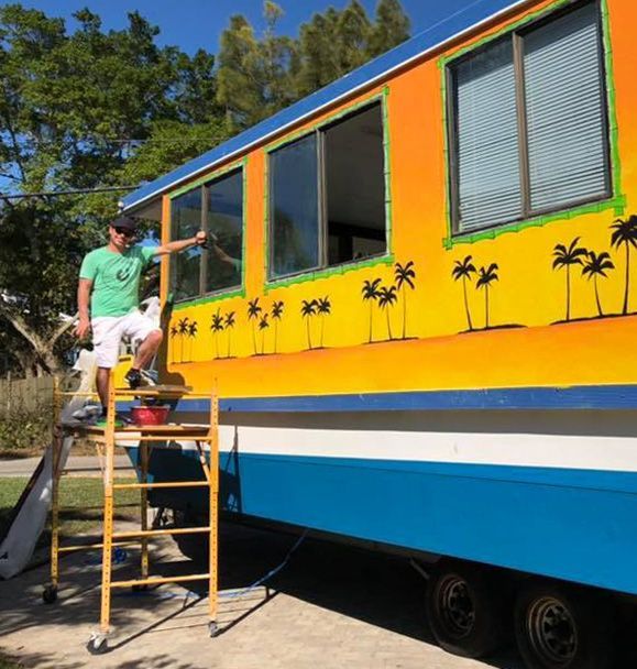 A man is standing on a ladder next to a colorful bus with palm trees painted on it