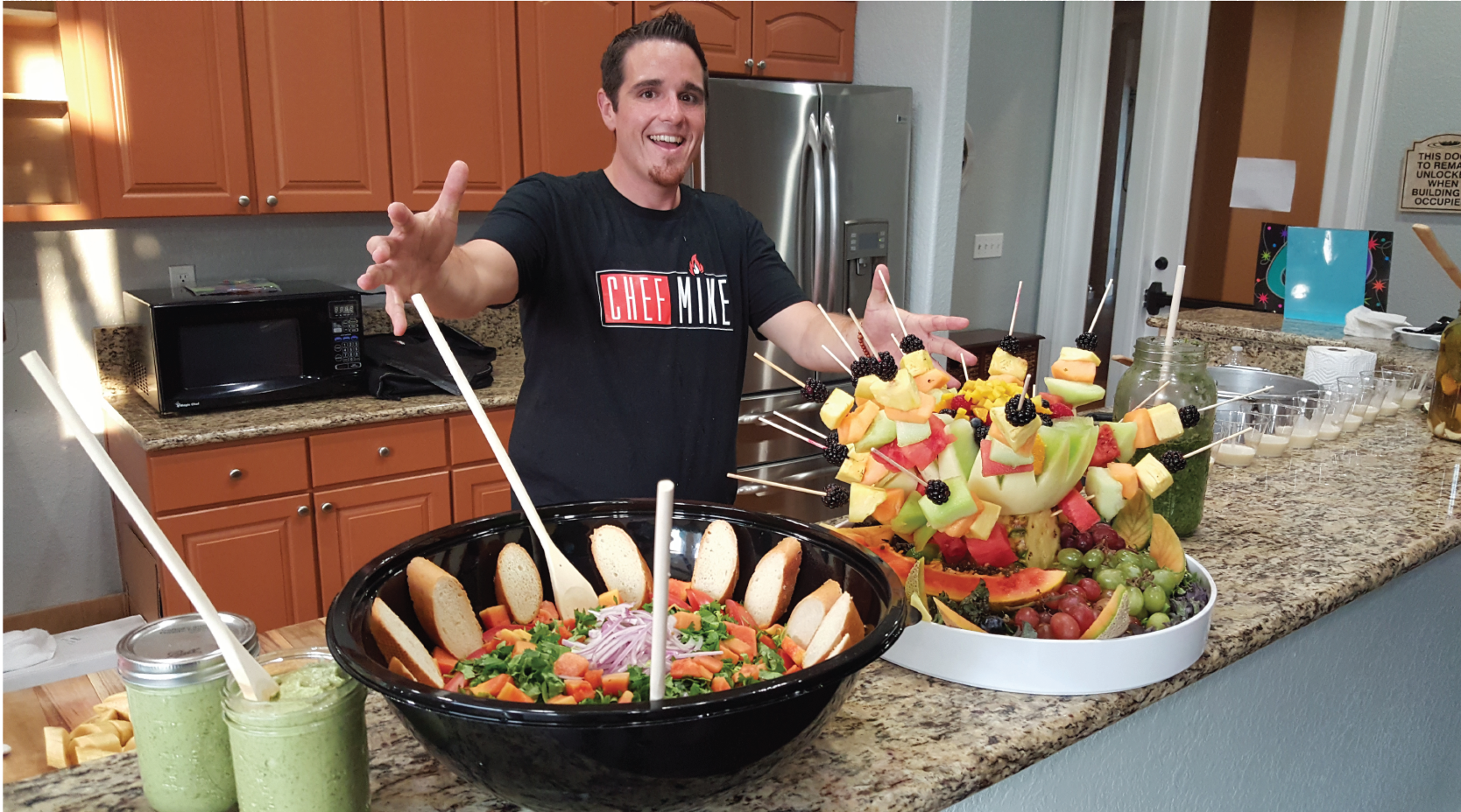 A man is standing in front of a bowl of fruit and smoothies.