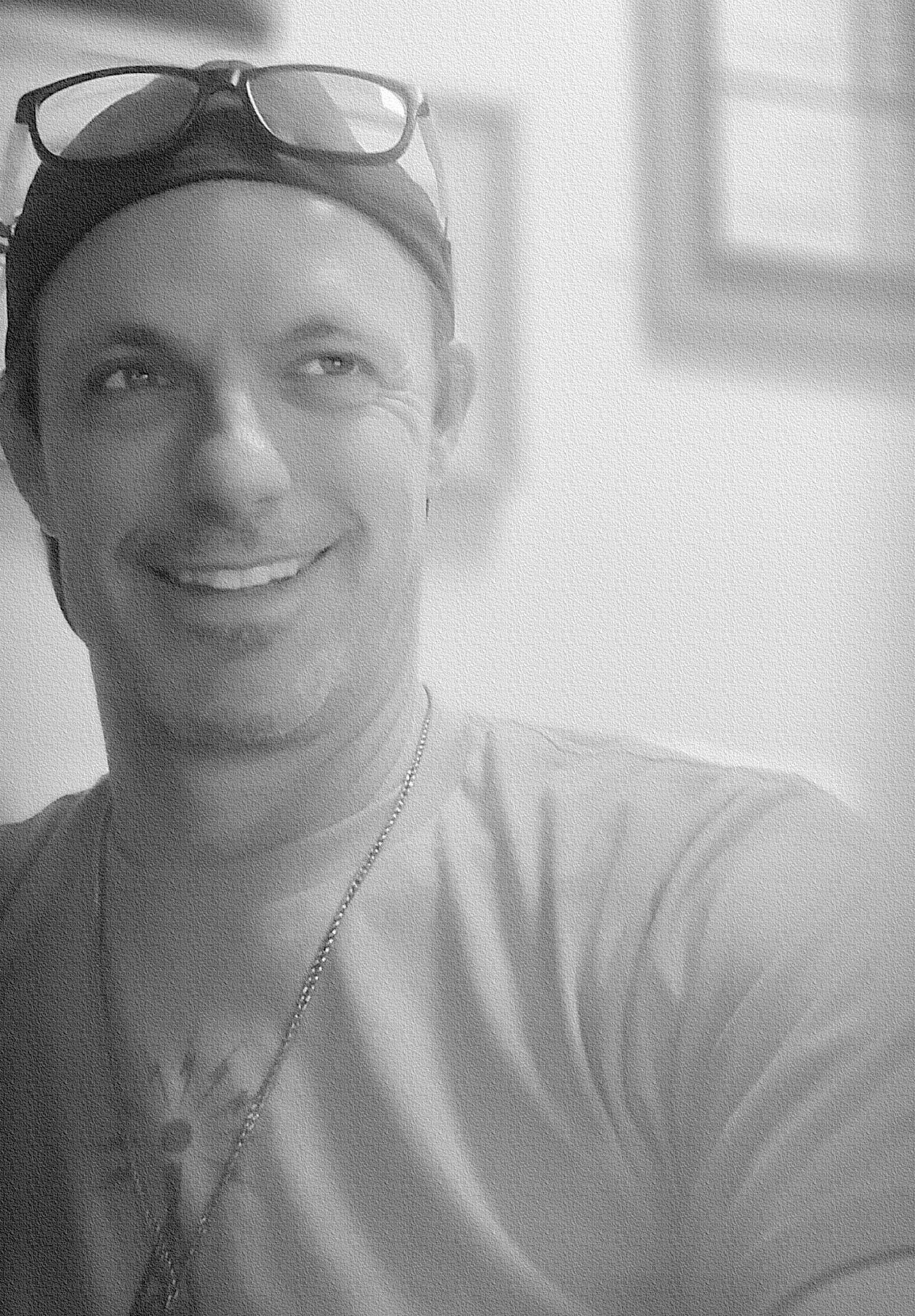 A man wearing sunglasses and a hat is smiling in a black and white photo.