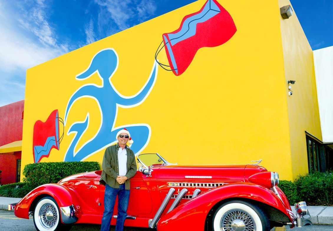 A man is standing next to a red car in front of a yellow building.