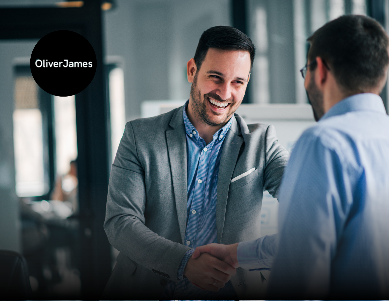 Two people in business attire smiling while shaking hands in an office setting with an Oliver James logo.