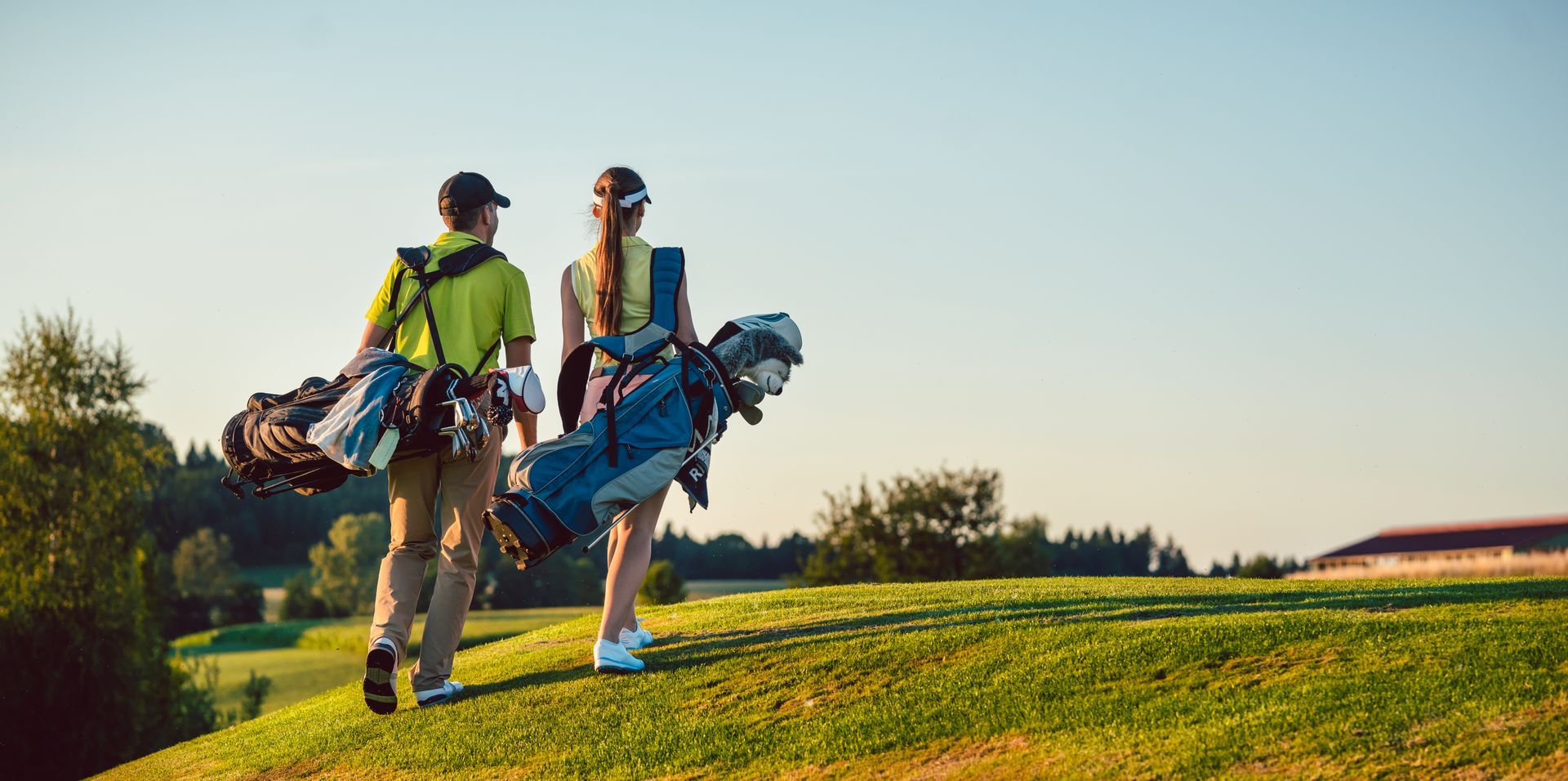 A man and woman walk across a golf course carrying golf bags. They are on a sunny day.