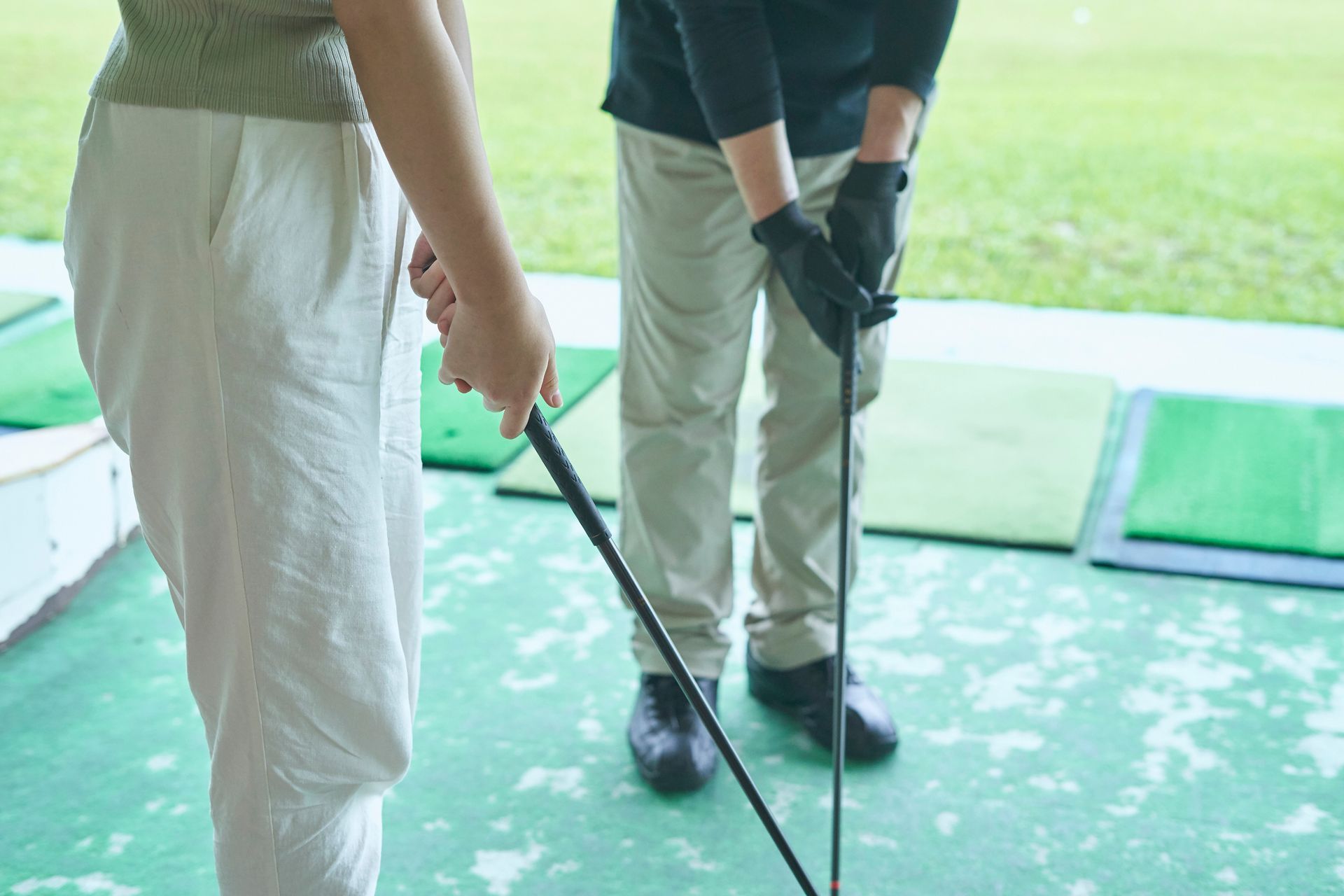 Two people practice golf at a driving range. One is holding a club and the other's arm is visible helping them.