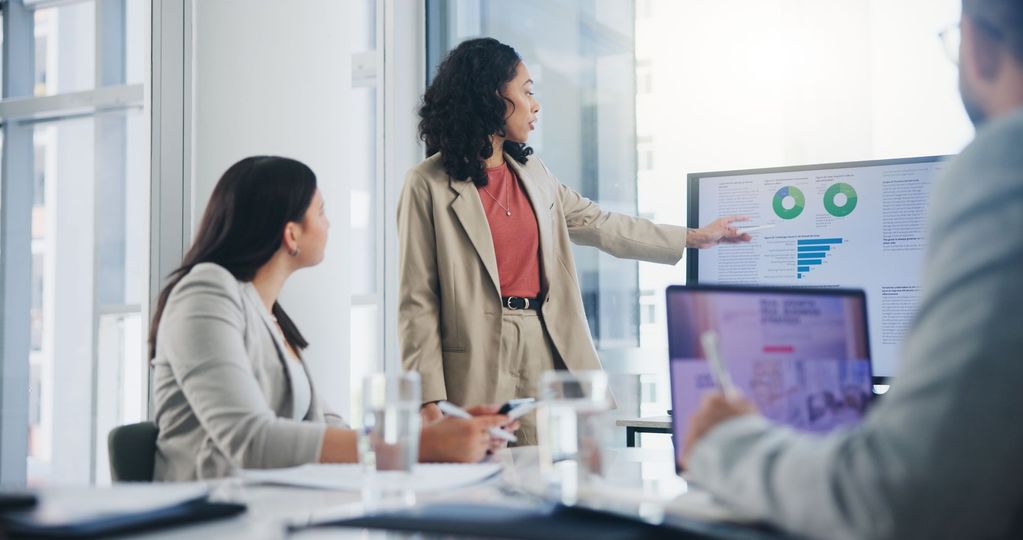Businesswoman presents data charts on screen to colleagues in an office meeting.