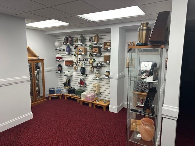 A display room with urns, jewelry, and keepsakes on shelves and in cabinets; red carpet and white walls.