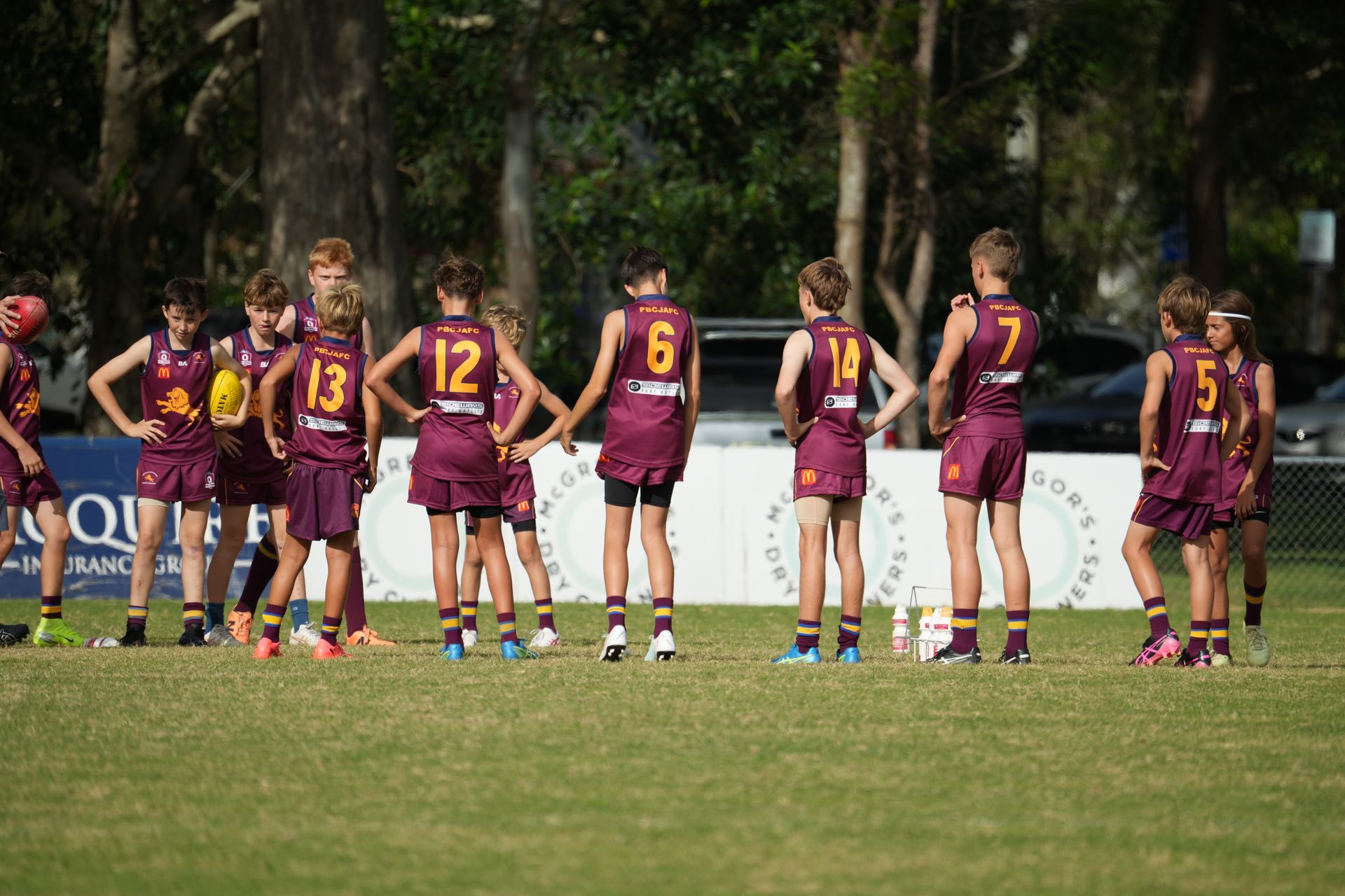 Maroon-and-gold athletes huddle on a grassy field, seen from behind near the sideline.